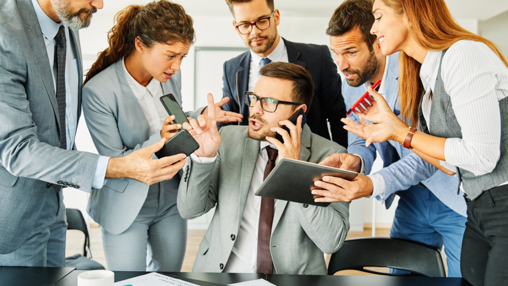 The image shows six business professionals surrounding a central figure who is on the phone and holding a tablet, appearing overwhelmed as others vie for his attention with phones and papers. 