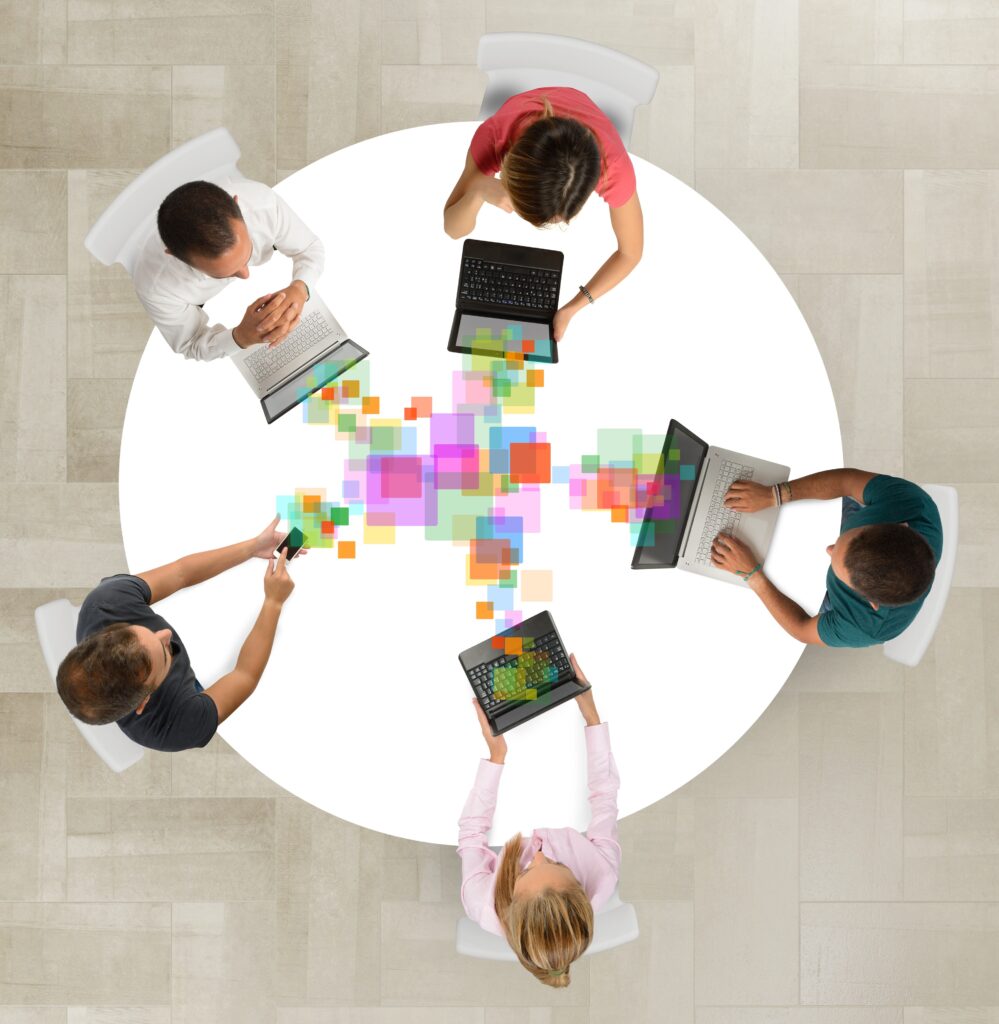 This image shows a top-down view of five people sitting around a circular table, each working on laptops or tablets. 