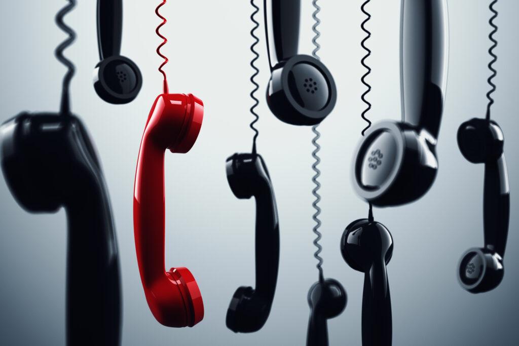 A red phone handset hanging among several black phone handsets, all with coiled cords