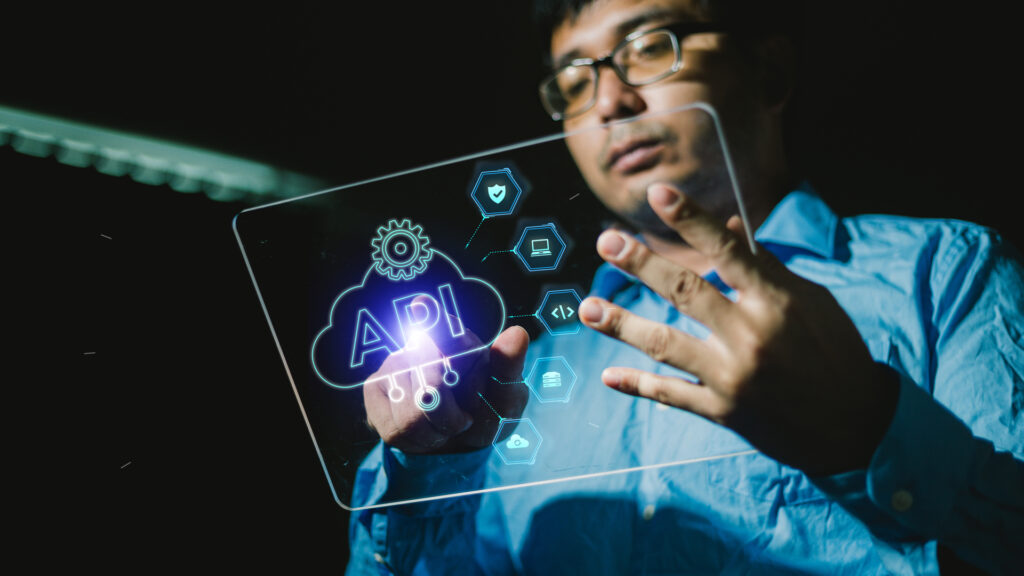 A person in a blue shirt interacts with a transparent futuristic touchscreen interface with the acronym ‘API’ prominently displayed in the center, surrounded by various icons representing settings, gears, and connectivity that symbolize API frameworks for software development.