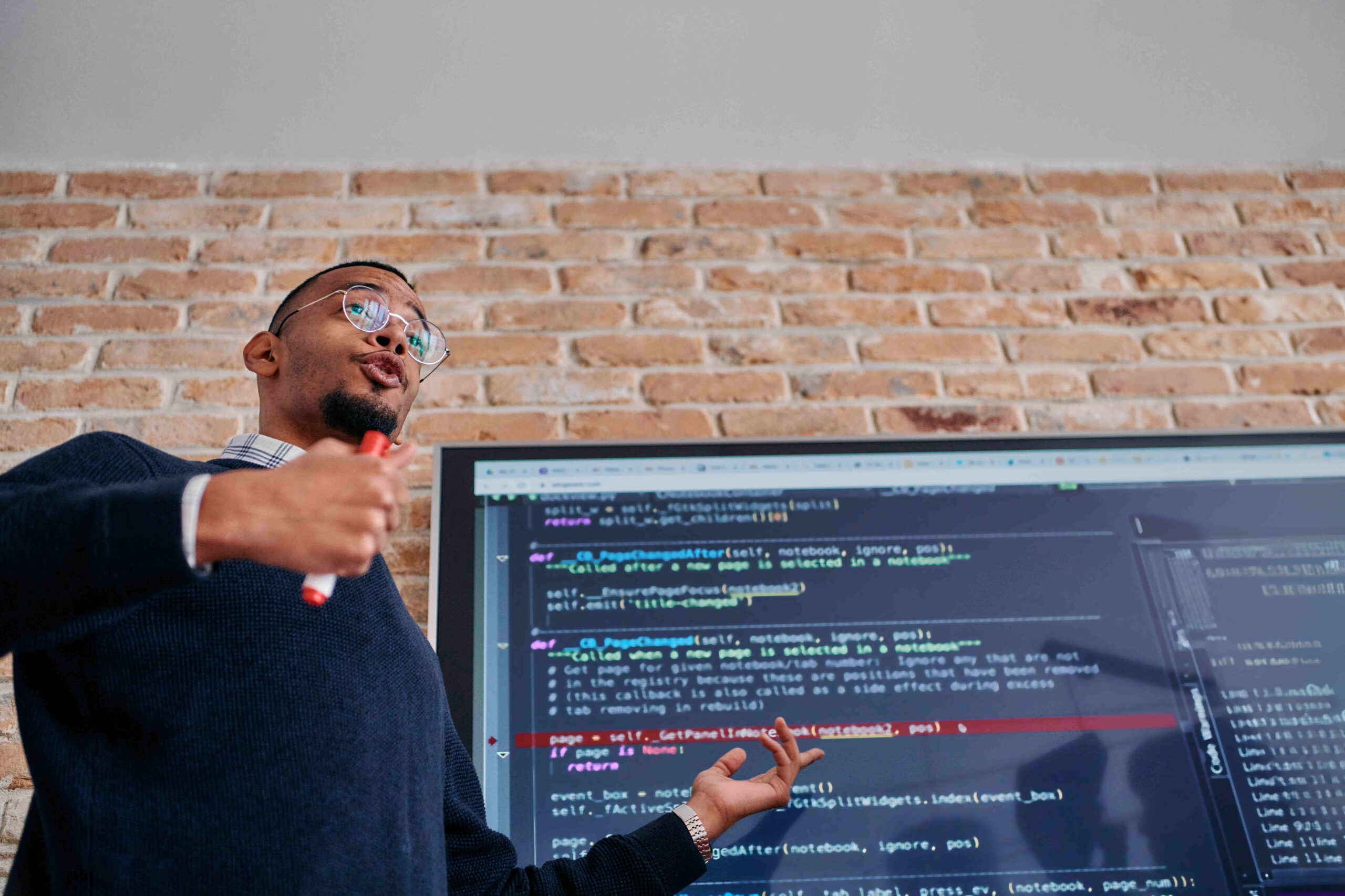 A person is standing in front of a large display screen with lines of code visible, likely discussing software development