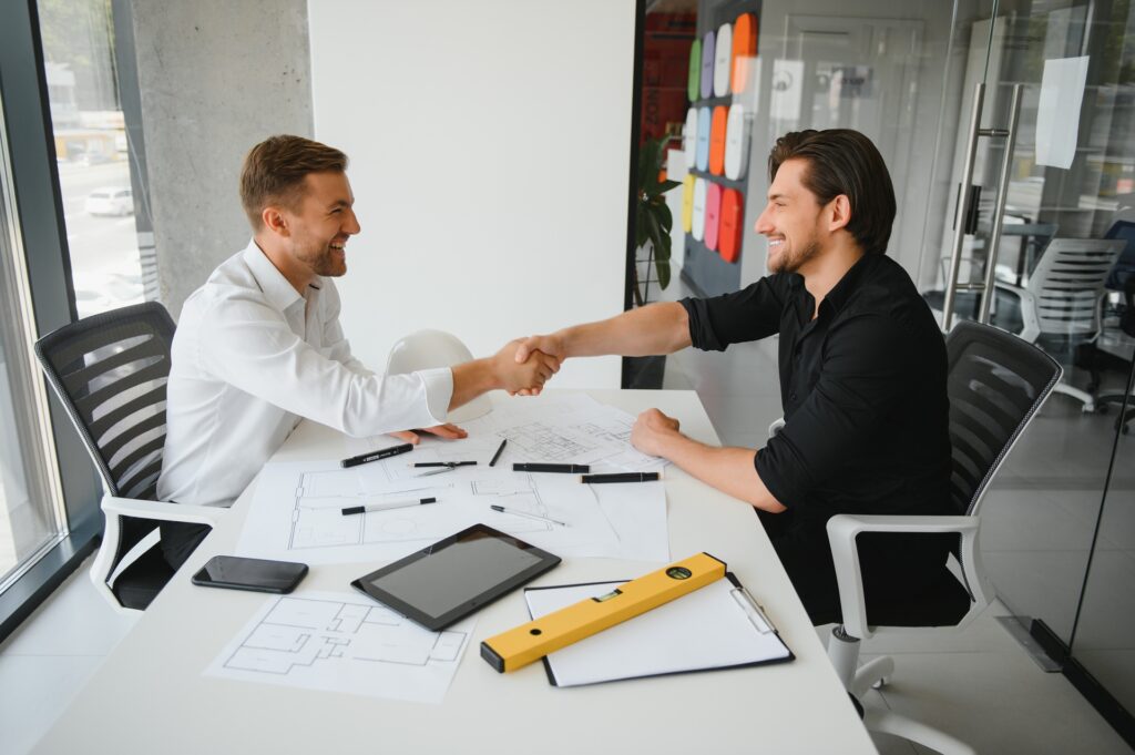 Two men smiling and shaking hands over a table filled with papers, a tablet, and drafting tools, symbolizing the agreement on a software purchase proposal in a modern office setting.