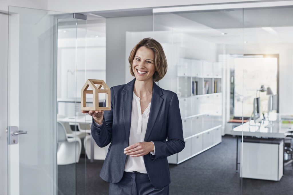 A professional woman in a modern office setting, holding a wooden model of a house. The image reflects concepts related to 'proptech' (property technology), combining real estate and technology, suggesting innovative solutions in property management, development, or real estate investment.