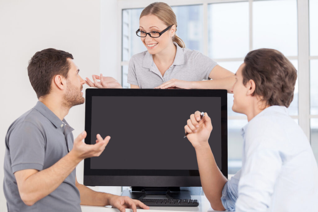 Three colleagues discussing a software purchase proposal in an office setting. A woman stands behind a large monitor, smiling and gesturing as she explains something to two seated men, who are engaged in the conversation.