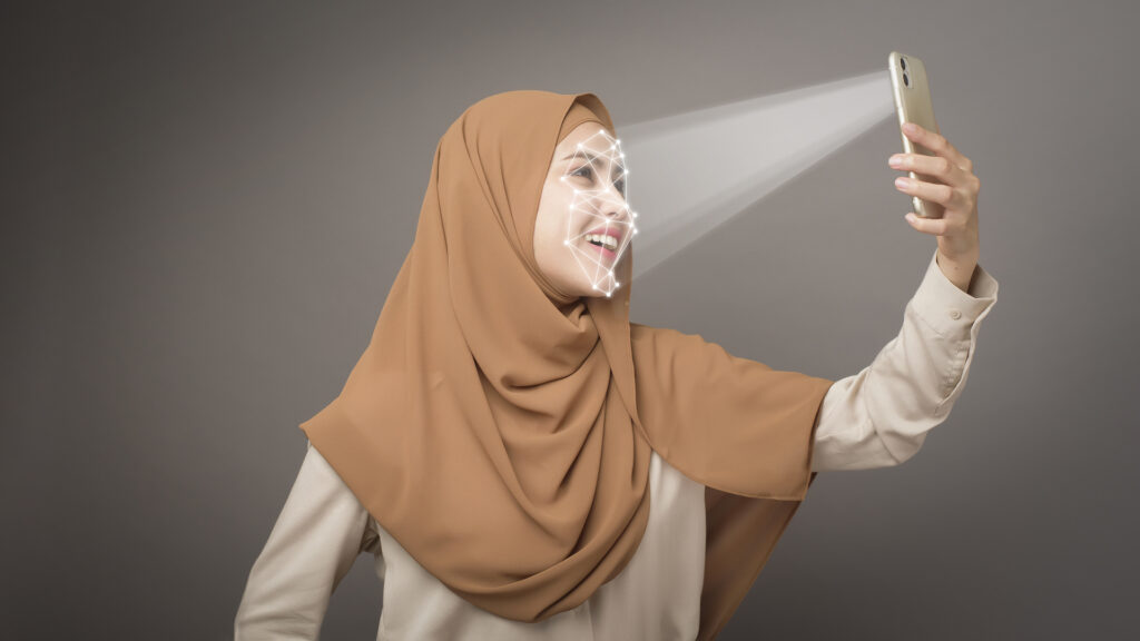 A woman wearing a hijab uses a smartphone for facial recognition, demonstrating advanced mobile software trends. The phone projects a light beam, creating a facial mapping grid on her smiling face. This image showcases the integration of AI-powered biometric technology in everyday devices, highlighting developments in mobile security, personalized user experiences, and inclusive software design that accommodates diverse user demographics.