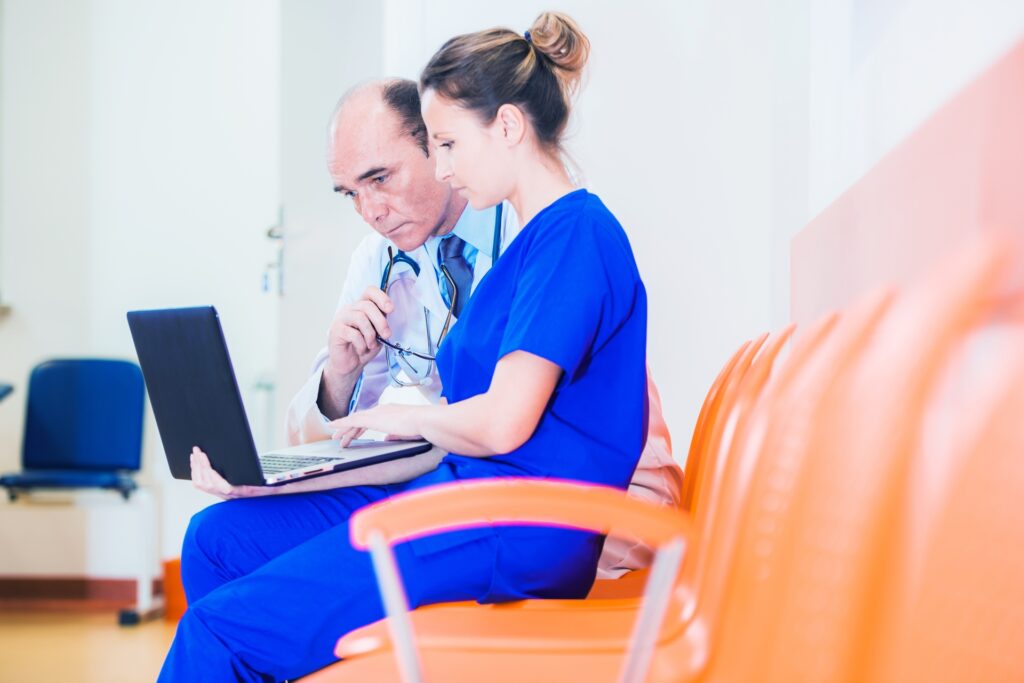 The image depicts two medical professionals - a doctor and a nurse or medical assistant - looking at a laptop together. They appear to be in a clinical setting, with the doctor wearing a white coat and stethoscope, and the other person in blue scrubs.