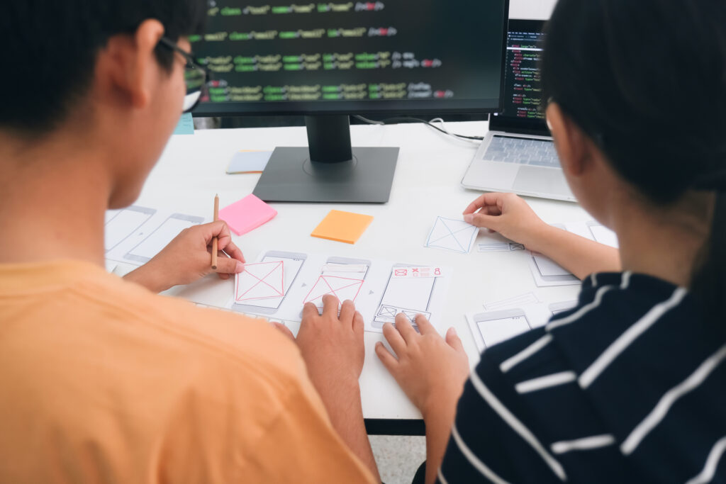 Two individuals engaged in experience engineering, one analyzing wireframes and user interface designs on paper, while the other codes on a computer with multiple lines of code visible on the screen.