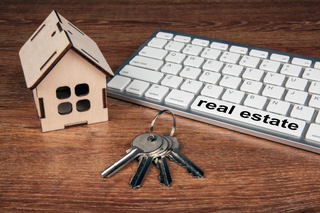 A wooden house model, keys, and a keyboard with the word "real estate" printed on it. Symbolic of using technology to find and manage properties.