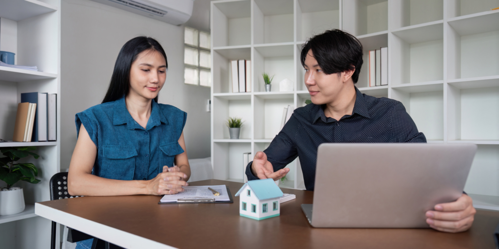 A customer and a professional are engaged in a discussion at an office desk, with a small model house on the table, symbolizing a customer relationship in real estate or financial services.