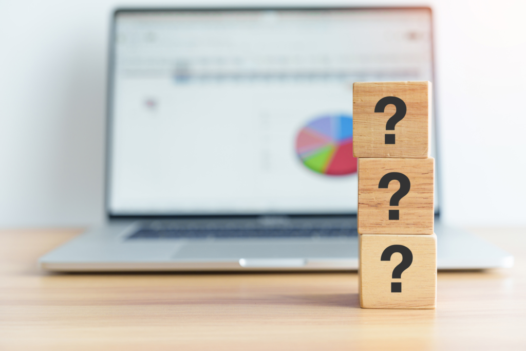 Three wooden blocks stacked on a desk, each with a question mark. A laptop with a chart visible in the background symbolizes the data-driven nature of product discovery.