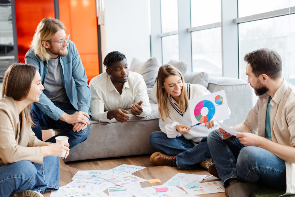 A diverse team of young professionals engaged in a product discovery session, analyzing data visualizations and charts spread across the floor while having an informal collaborative discussion in a modern office space