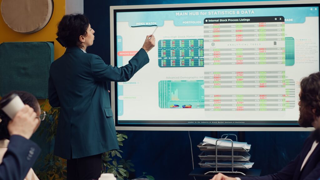 A professional presents marketing operations data on a large interactive screen, showing stock market listings, demographics, and analytical tools, in a business meeting setting.