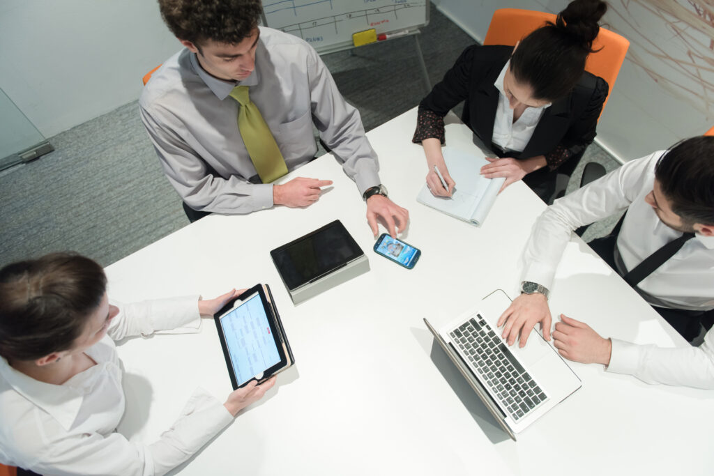 A team of four professionals collaborates during the project discovery phase, utilizing various devices such as tablets, laptops, and smartphones to share and discuss information.