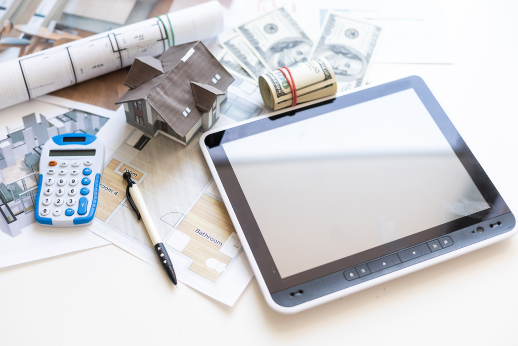 Real estate software concept with a tablet, architectural plans, a model house, calculator, pen, and rolled-up dollar bills, illustrating tools used in property planning and financial calculations.