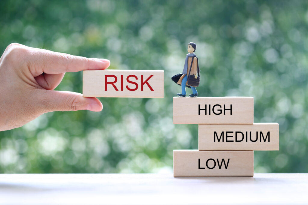 A hand holds a block labeled "RISK" in front of a figure standing on blocks labeled "HIGH," "MEDIUM," and "LOW," symbolizing risk assessment levels.