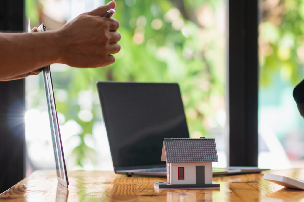 A workspace with a laptop and a small model house on a wooden table, with a person holding a tablet in the background. The scene suggests the integration of technology in SaaS marketing websites, focusing on real estate.