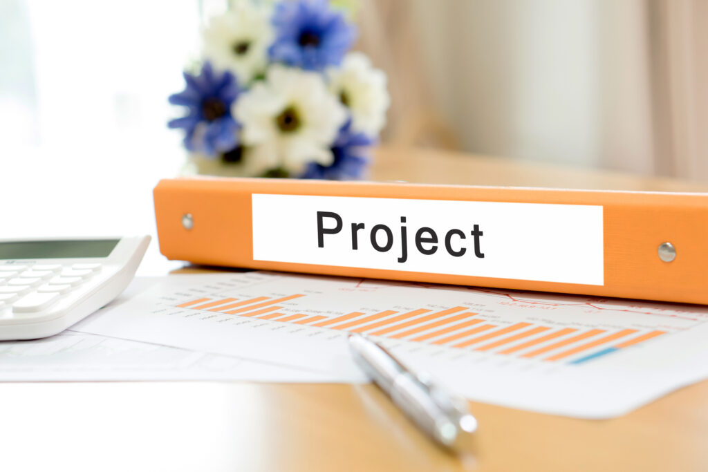 An orange binder labeled 'Project' sits on a desk with a bar chart, pen, and calculator, representing project deliverables and planning