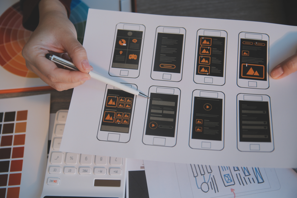 The image shows a person holding a sheet with wireframes and prototypes of a mobile app, highlighting the design process from initial layout (wireframe) to detailed visual representation (prototype).