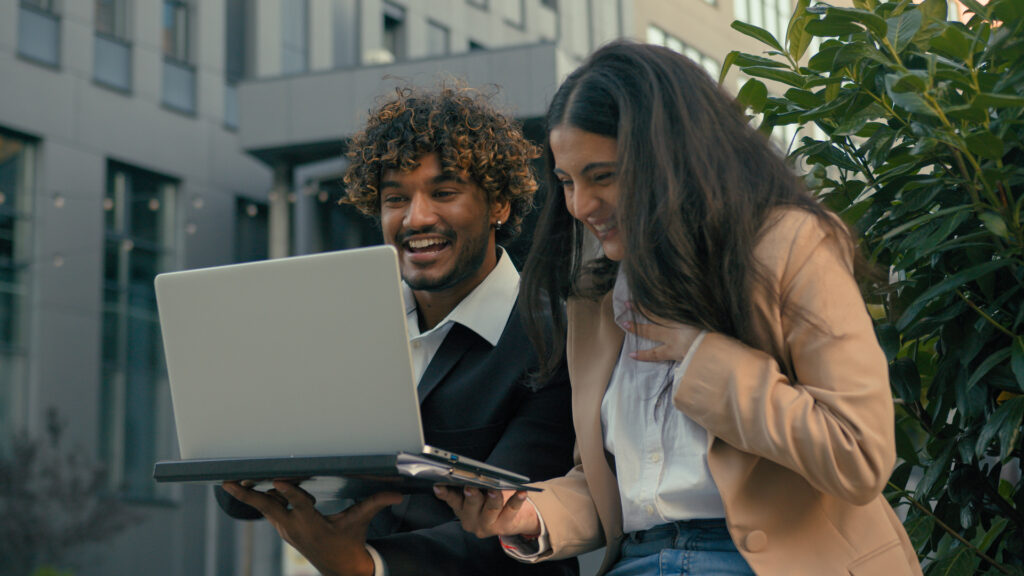 A joyful pair explores product discovery techniques on a laptop, laughing together outdoors. They are engaged and enthusiastic, reflecting the excitement of finding innovative solutions.