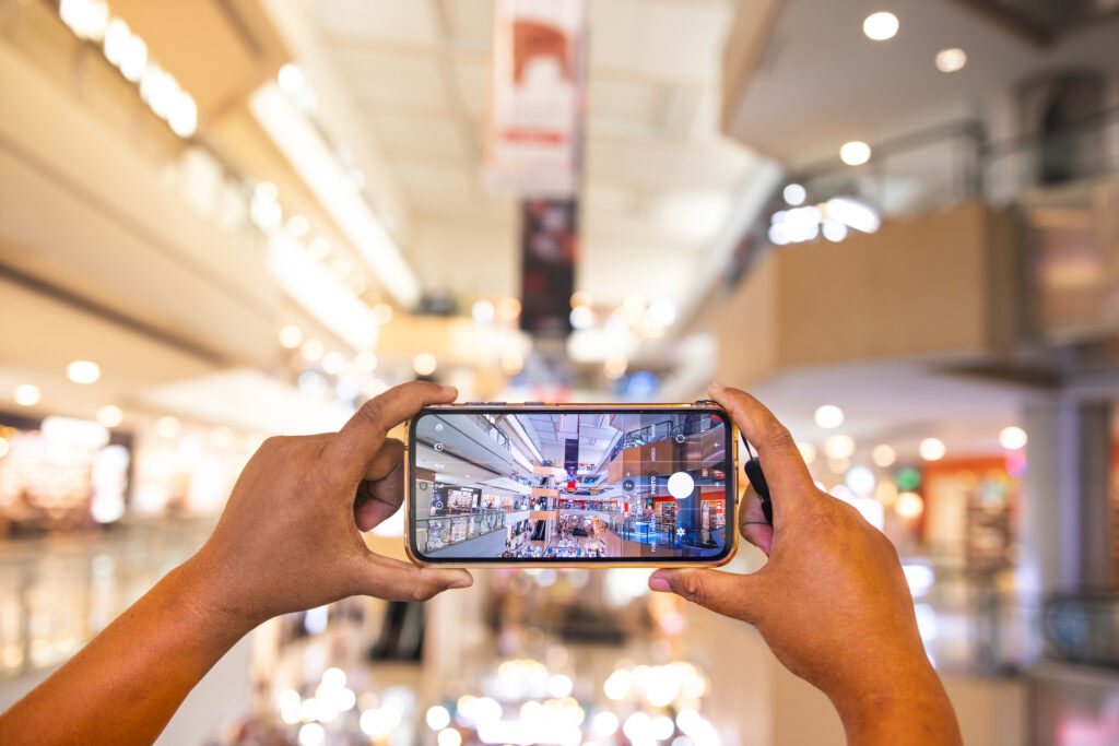 A person captures a photo of a bustling shopping mall with their smartphone. This visualizes the use of mobile devices for market research and product discovery.