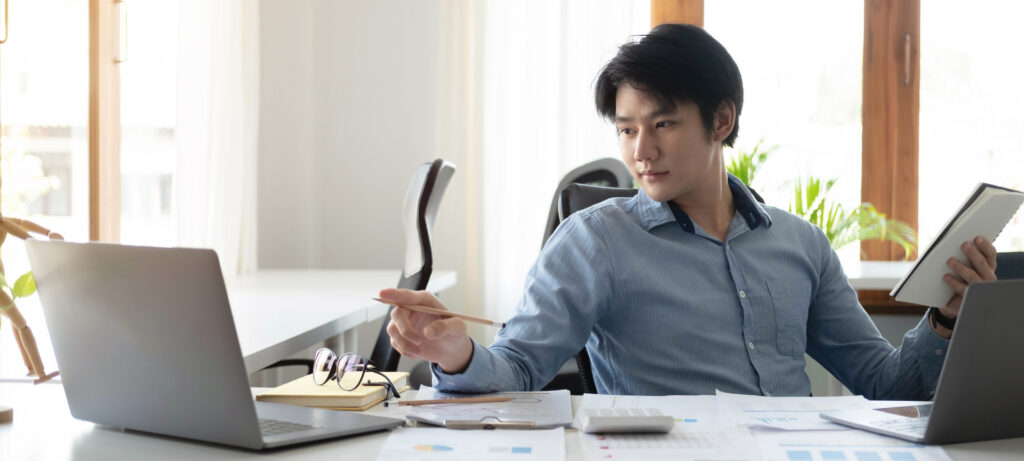 A young professional analyzes data on a laptop, holding a notebook and a pencil, suggesting a focus on precision and strategic planning in target marketing.