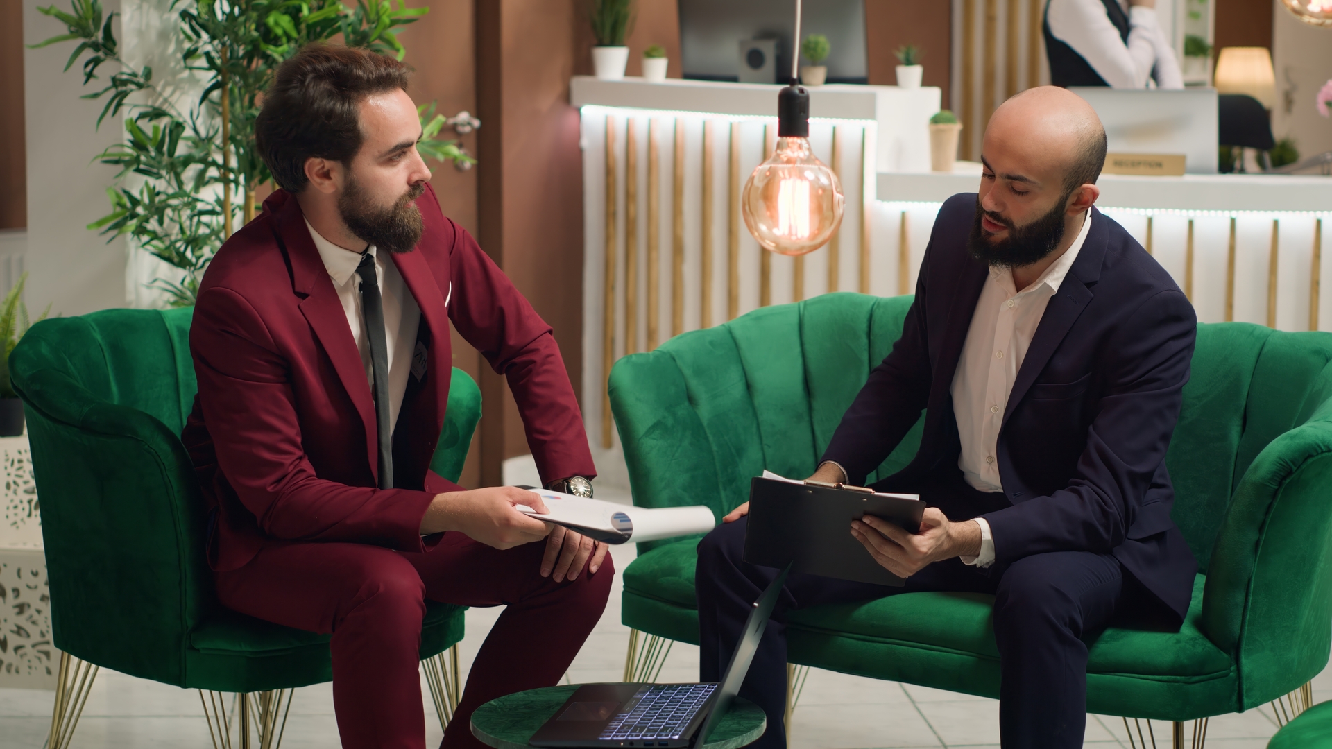 Two individuals in suits, one holding a document and the other using a tablet, sit on a green sofa. The setting suggests a discussion or review of “Regulation F.”