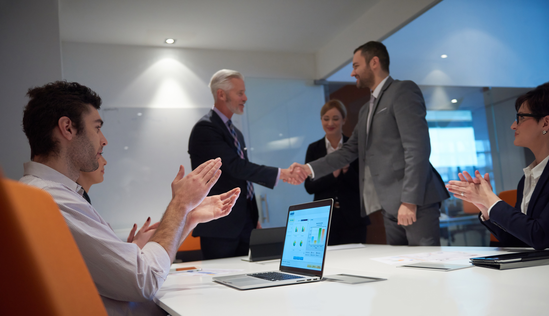 A professional meeting where team members applaud as two individuals shake hands, with a laptop displaying software analytics on the table, highlighting the success of a software demo presentation.