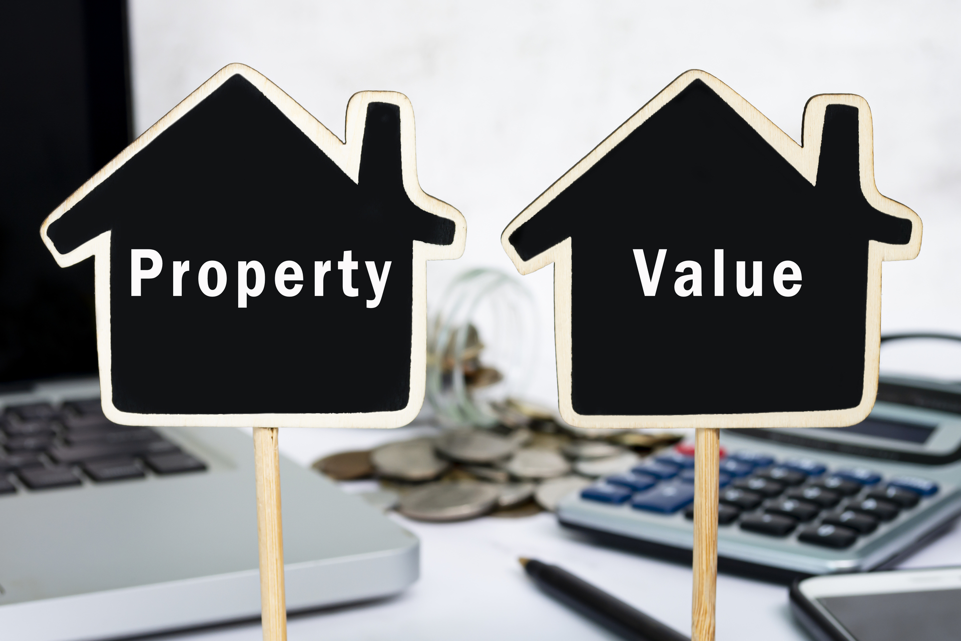 Two house-shaped signs labeled "Property" and "Value" sit on a desk with a laptop, calculator, and coins. This image could represent the importance of understanding property values when targeting potential buyers or sellers.